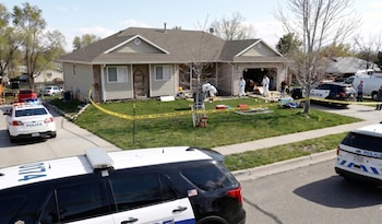 Casa residencial con césped acordonado con cinta amarilla "NO CRUZAR". Personal forense en trajes blancos trabaja en la escena. Dos coches de policía están en la calle