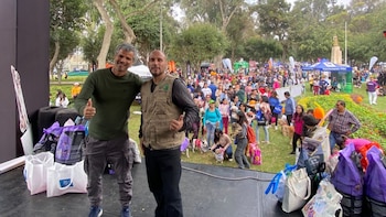 Dos hombres en un escenario saludan, rodeados de bolsas. Detrás, una multitud de personas con perros en un parque con árboles y tiendas