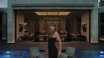Taylor Swift con vestido negro frente a una piscina iluminada, dos sillas de piedra y una edificación de hormigón al fondo.