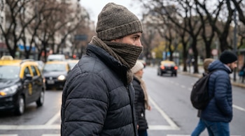 Plano corto de un hombre de perfil con gorro de lana y bufanda gruesa que cubre su boca, vistiendo una campera acolchada, cruzando una avenida en Buenos Aires.