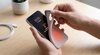 Manos retirando la funda de un smartphone oscuro. La pantalla muestra "1% Batería" y una alerta de "Sobrecalentamiento" con un termómetro rojo. Se ve un cable de carga.