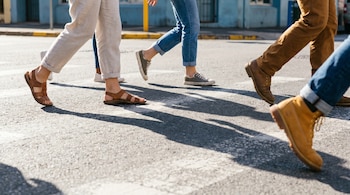 Primer plano de piernas y pies de varias personas caminando por un paso de cebra en una calle con asfalto gris y sombras alargadas. Se ven sandalias, zapatillas y botas.