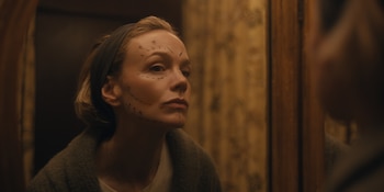 Carey Mulligan mira su reflejo en un espejo. Su rostro tiene líneas negras y flechas dibujadas. Lleva un pañuelo en la cabeza y un suéter oscuro