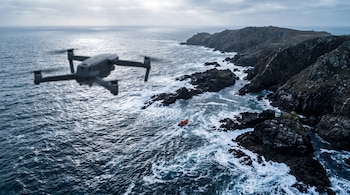 Un dron gris vuela en primer plano sobre un mar de olas con espuma, una embarcación naranja y acantilados rocosos.