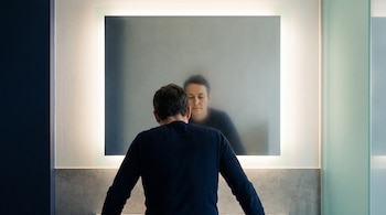 Persona adulta de espaldas con postura tensa frente a un espejo iluminado. El reflejo del rostro en el espejo se ve borroso y sereno.