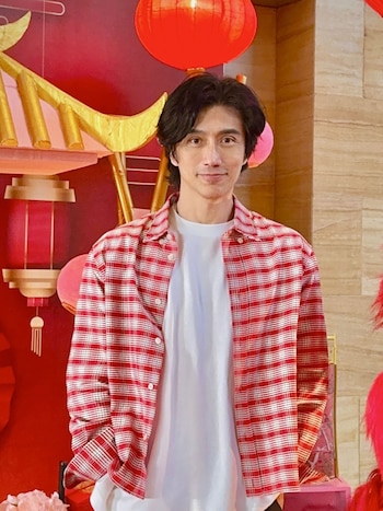 Un hombre con camisa a cuadros roja y camiseta blanca se encuentra frente a faroles rojos y estructuras doradas