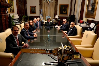 Ocho personas, siete hombres y una mujer, sentadas alrededor de una mesa. Una motosierra dorada y negra está en el primer plano. Cuadros y bandera argentina al fondo