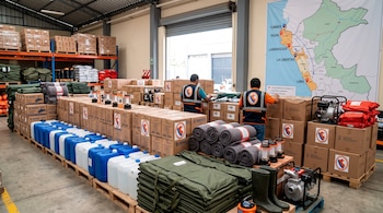 Palés con cajas de cartón con el logo de Indeci, bidones de agua, mantas, motobombas y dos trabajadores frente a un mapa mural de Perú.