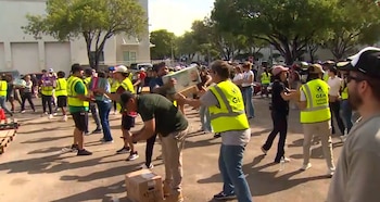 Grupo de personas en una zona exterior pasando cajas. Varias personas visten chalecos reflectantes amarillos. Hay palés de madera, árboles y edificios al fondo