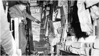 Un hombre con sombrero y traje, de perfil, se apoya en un barandal y señala hacia un estrecho pasillo abarrotado de pilas de periódicos, cajas y objetos, en blanco y negro