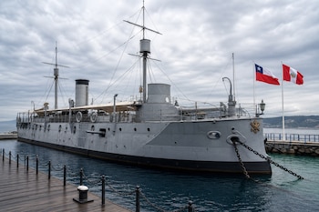 Buque de guerra gris amarrado a un muelle de madera junto a dos mástiles con las banderas de Chile y Perú bajo un cielo nublado.