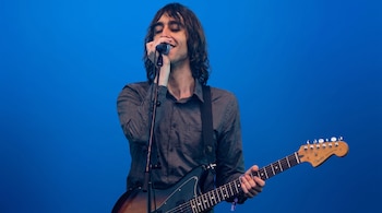 Hombre de cabello largo canta en un micrófono y toca una guitarra eléctrica con correa. Viste camisa oscura y el fondo es azul liso.