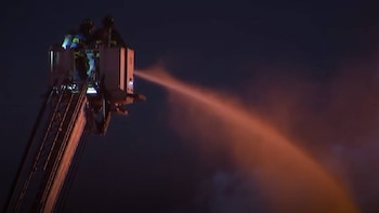 Vista nocturna de dos bomberos en la cesta de una escalera de camión, proyectando un chorro de agua o espuma con un brillo rojizo contra un fondo oscuro