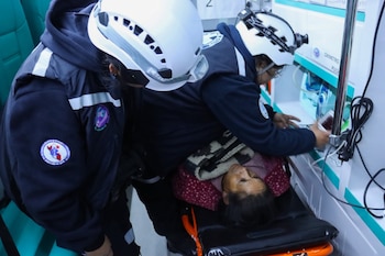 Dos rescatistas con uniformes y cascos blancos atienden a una persona en una camilla dentro de una ambulancia con equipo médico