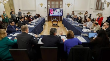 Sala de reuniones formal con dos largas mesas en forma de U, donde varias personas están sentadas. Una pantalla central muestra una videollamada con dos hombres
