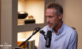 Mark Hyman viste una camisa azul frente a un micrófono de estudio en un espacio iluminado con estantes al fondo