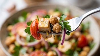 Tenedor con un cubo de pollo, garbanzos, perejil, tomate y cebolla morada con gotas de aderezo, frente a un cuenco de ensalada.