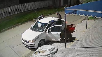 Vista aérea nocturna de un coche blanco estacionado en la calle, con varias personas, algunas subiendo al vehículo. Un toldo azul a la derecha