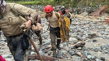 Un grupo de rescatistas y civiles con ropa embarrada caminan por un lecho de río rocoso. Un rescatista carga una cuerda amarilla y un pico