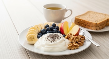 Plato con yogur, semillas de chía, rodajas de banana, arándanos, manzana y nueces. Al fondo, taza de té verde y pan integral en mesa de madera clara.