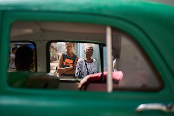 Dos mujeres, una joven y otra anciana, se ven a través de la ventanilla de un coche verde, con una calle borrosa al fondo bajo la luz del día