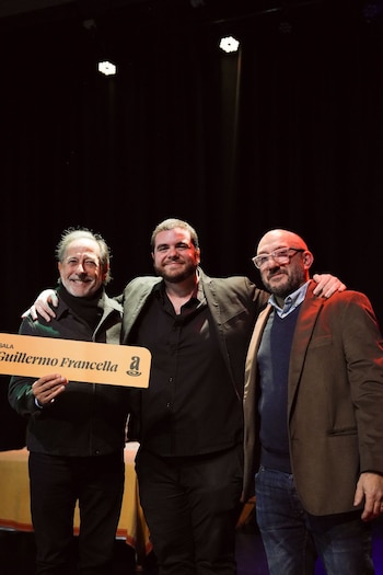 Tres hombres sonríen y posan juntos en un escenario oscuro con focos de luz; uno de ellos sostiene un cartel que dice "SALA Guillermo Francella"