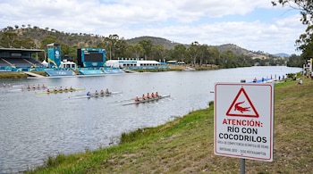Un cartel de advertencia sobre cocodrilos se encuentra junto a un río con embarcaciones de remo, tribunas y una pantalla gigante.