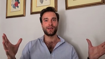 Primer plano de un hombre con barba, camisa de rayas azul clara, gesticulando con ambas manos. Detrás, una pared blanca con dos cuadros botánicos