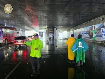 Agente de tráfico con impermeable amarillo dirige el tráfico en un paso a desnivel inundado. Al fondo, dos personas con impermeables observan el nivel del agua