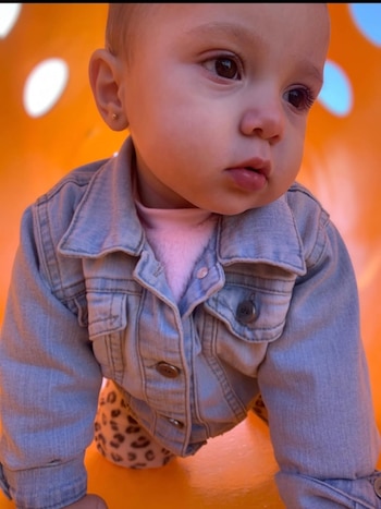 Primer plano de una niña pequeña gateando sobre una superficie naranja, vestida con una chaqueta vaquera azul claro y top rosa. Mira hacia la derecha