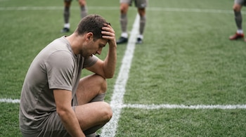 Hombre futbolista de rodillas con la mano en la frente, vistiendo camiseta gris, sobre césped verde con líneas blancas. Otros jugadores borrosos al fondo.