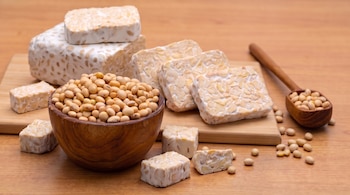 Porciones y bloques de tempeh junto a un cuenco de madera y una cuchara con granos de soja sobre una mesa de madera.