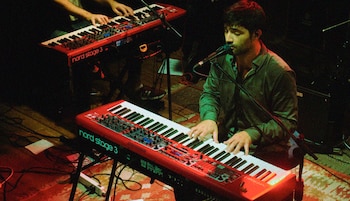 Primer plano de un músico tocando un teclado Nord Stage 3 rojo y cantando. Otro músico y su teclado rojo son visibles atrás, en un escenario con luces cálidas