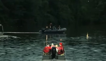 Dos botes navegan en un río oscuro, con personas a bordo. El bote más cercano muestra tres individuos con chalecos salvavidas rojos. Un bosque oscuro bordea el río
