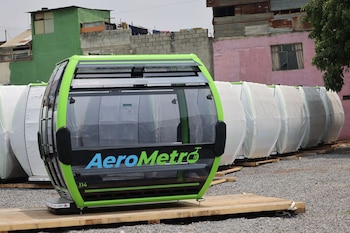 Una cabina verde y negra de AeroMetro, con el número 114, sobre una tarima de madera en un suelo de grava. Varias cabinas cubiertas y edificios de fondo