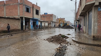 Calle cubierta de agua marrón y lodo frente a viviendas de ladrillo, con peatones en las aceras y una excavadora amarilla al fondo.