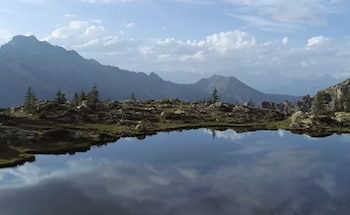 Un lago de montaña (tarn) con la superficie oscura que refleja el cielo y las montañas. En primer plano, rocas y hierba; montañas escarpadas al fondo