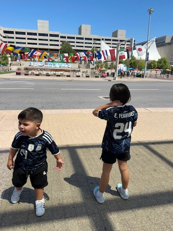 Dos niños con camisetas de fútbol con el número 24. Uno tiene el nombre "E. FERNANDEZ" en la espalda. Edificio y banderas internacionales de fondo