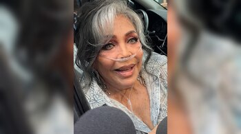 Mujer adulta mayor con cabello gris y negro, cánula nasal de oxígeno en la nariz, aretes largos y una blusa estampada, sentada en un automóvil.