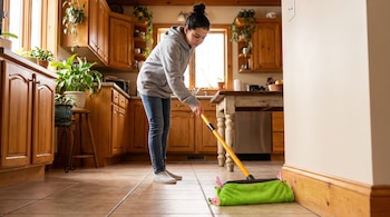 Joven de pie con sudadera gris y jeans limpia un zócalo de madera en una cocina con azulejos. Usa una escoba amarilla con paño verde.
