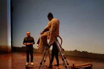 Un actor con vestuario estampado sobre una escalera de aluminio, una asistente detrás y un hombre de pie en el escenario