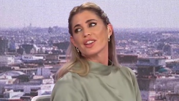 Primer plano de Pamela David, mujer rubia con cabello recogido en trenzas y blusa verde oliva, mirando hacia un lado con un fondo urbano difuminado