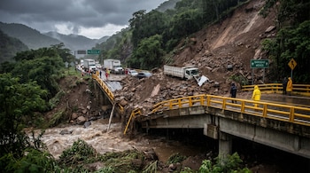 Carretera montañosa dañada por un derrumbe masivo y un puente colapsado sobre un río turbio. Se ven vehículos varados y personas con impermeables bajo un cielo nublado.