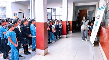 Varias estudiantes uniformadas escuchan a una mujer con micrófono y chaleco frente a un panel con el emblema de la Sucamec