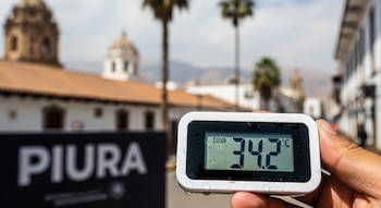 Una mano sostiene un termómetro digital blanco con gotas de agua que muestra 34.2 °C, con un cartel borroso con la palabra Piura y edificios al fondo.