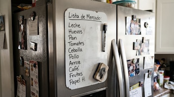 Una lista de mercado escrita a mano en una pizarra magnética en la puerta de un refrigerador de acero inoxidable. Un marcador y un borrador están pegados al tablero.