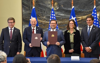 Cinco funcionarios sonríen mientras dos hombres al centro sostienen documentos. Detrás, banderas de Chile y una azul con estrellas blancas