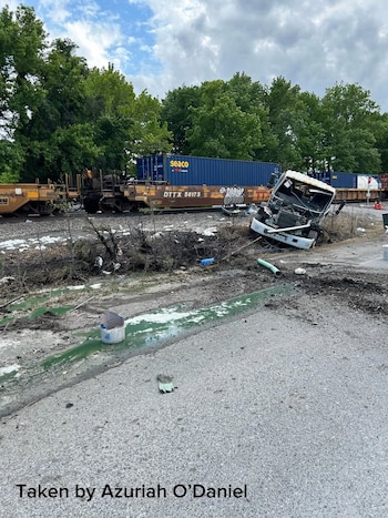 Un tren de carga con contenedores azules y un camión blanco destrozado en un paso a nivel. Hay escombros y un derrame de líquido verde en el suelo