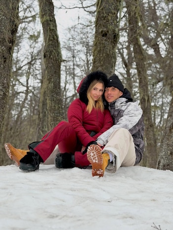 Un hombre y una mujer vestidos con ropa de abrigo invernal posan sentados sobre la nieve rodeados de árboles