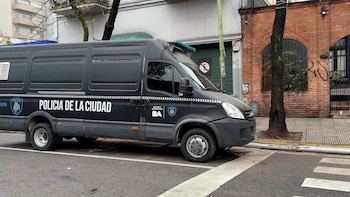 Furgón negro de la Policía de la Ciudad estacionado en la calle junto a la acera, árboles y fachadas de edificios al fondo